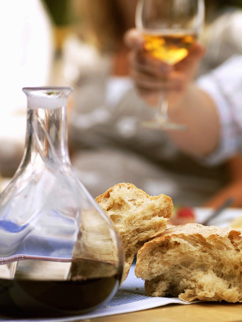 Rotwein und Brot auf Tisch, Hand hält Weinglas im Hintergrund