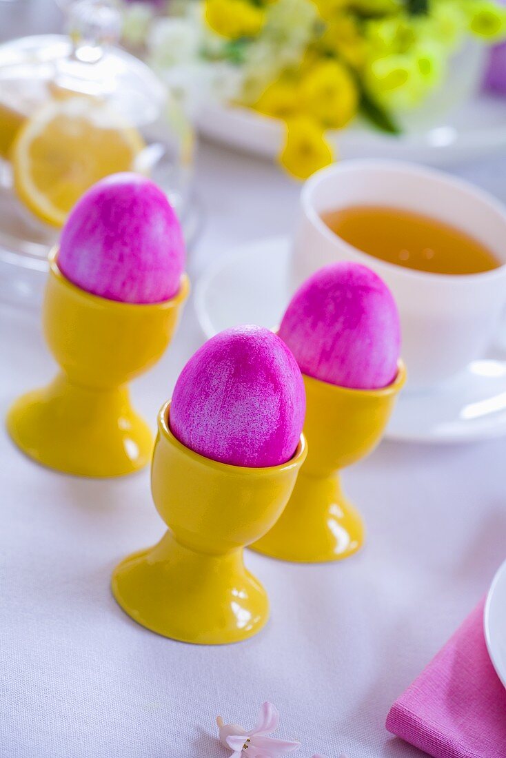 Pinkfarbene Ostereier in Eierbechern, dahinter Teetasse