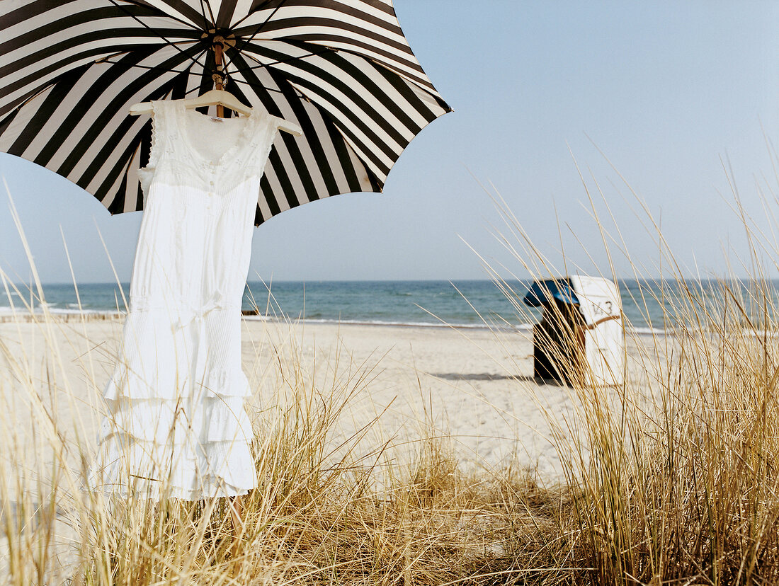 Rüschenkleid am Kleiderbügel unter Sonnenschirm am Strand, Strandkorb