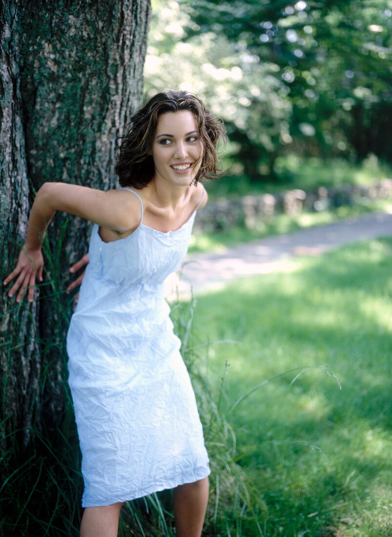 Cheerful woman wearing blue dress leaning against tree in park, smiling and looking away