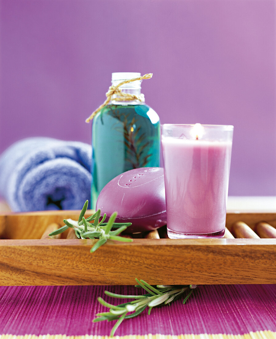 Aroma candle, soap and rosemary on wooden tray with towel and oil bottle in background