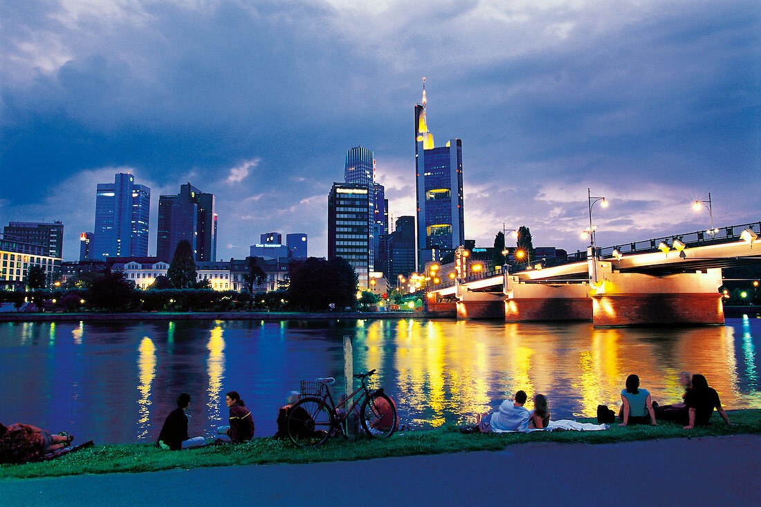People sitting near main river bank with skyline and illuminated cityscape