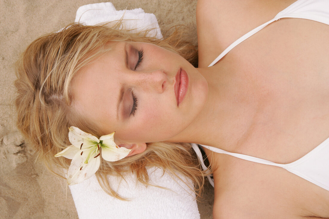 Pretty woman wearing white bikini lying on sand with towel under her head, eyes closed