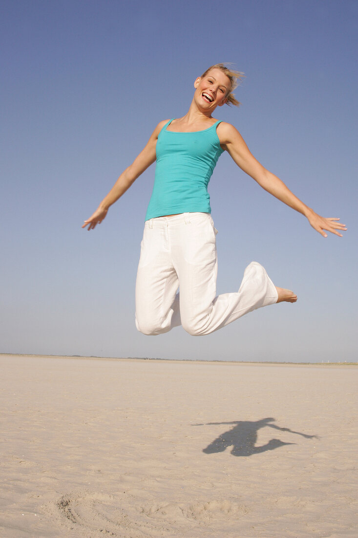 Pretty woman in blue top and white pants jumping on beach with arms outstretched, smiling