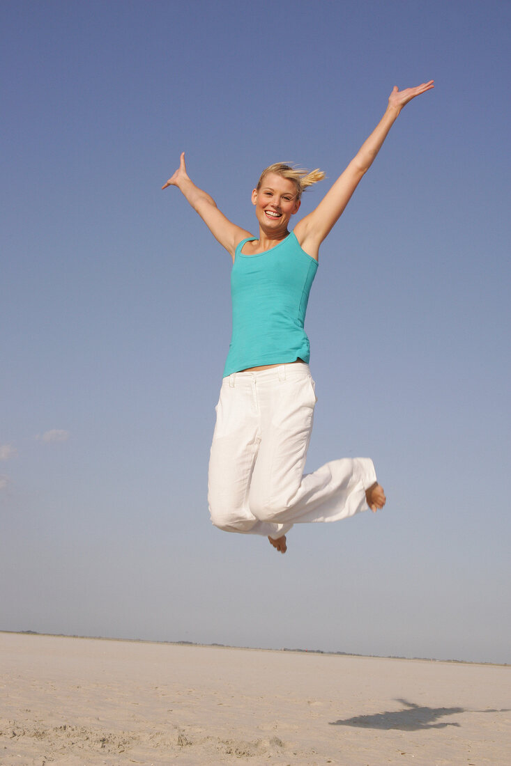 Pretty woman in blue top and white pants jumping on beach with arms outstretched, smiling