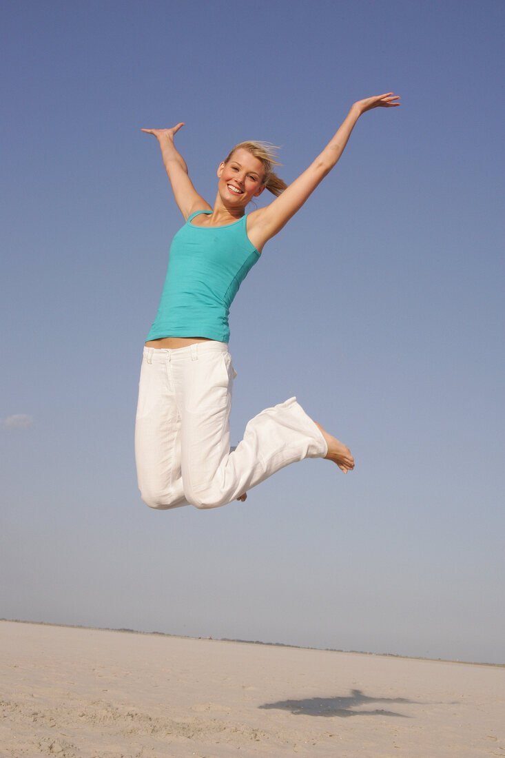 Pretty woman in blue top and white pants jumping on beach with arms outstretched, smiling