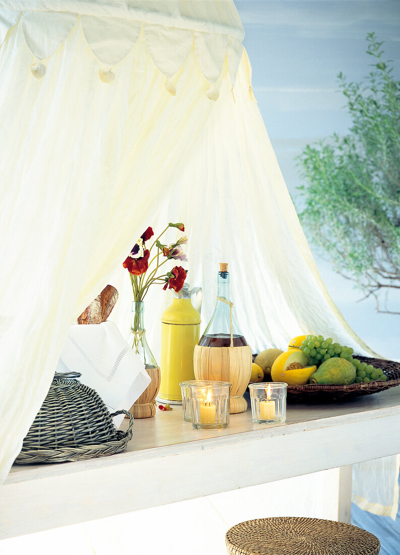 Mosquito net over table with fruit wine, candles, wicker basket and flower pot