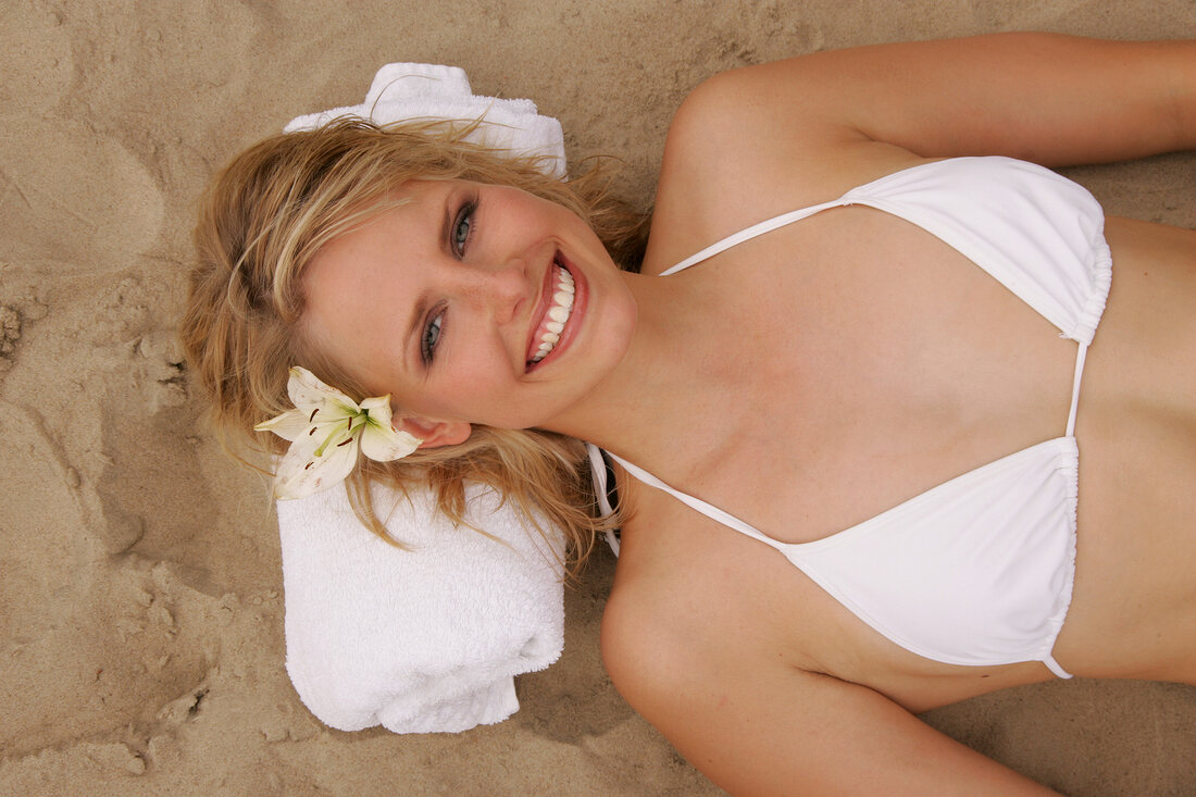 Overhead view of pretty woman in white bikini with flower in hair lying on sand, smiling