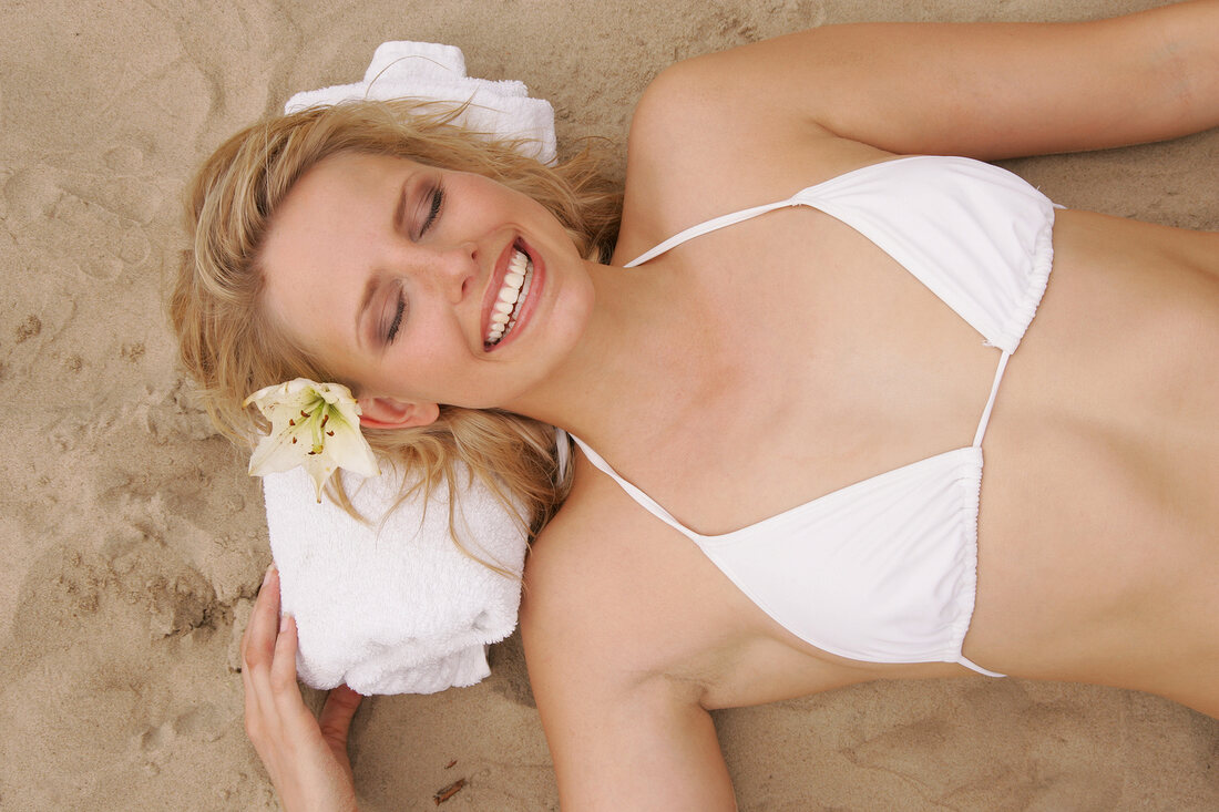 Overhead view of pretty woman in white bikini lying on sand, smiling with eyes closed