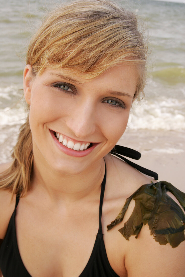 Portrait of beautiful woman in black bikini top sitting with seaweed on shoulder, smiling