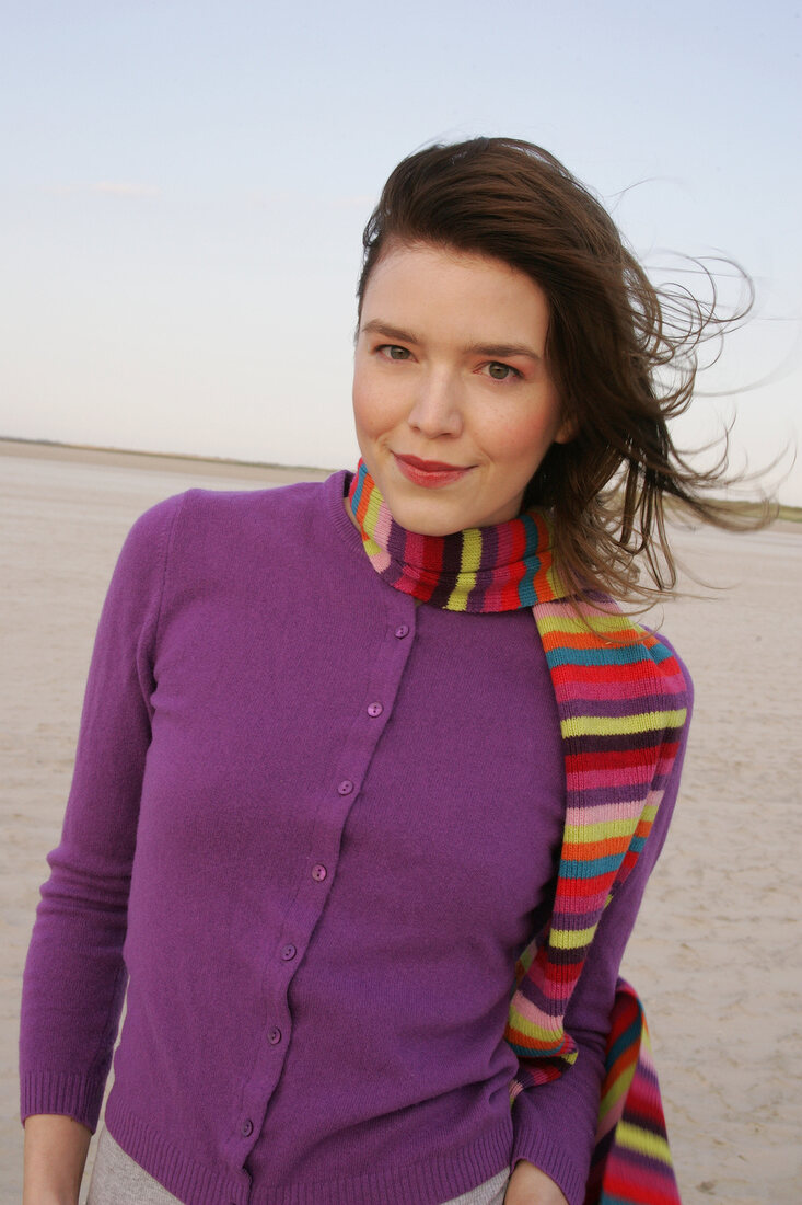 Pretty blonde woman wearing purple sweater and striped scarf standing on beach, smiling