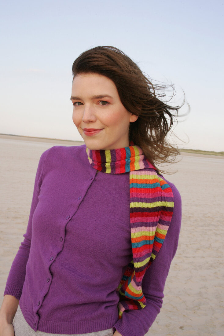 Pretty blonde woman wearing purple sweater and striped scarf standing on beach, smiling