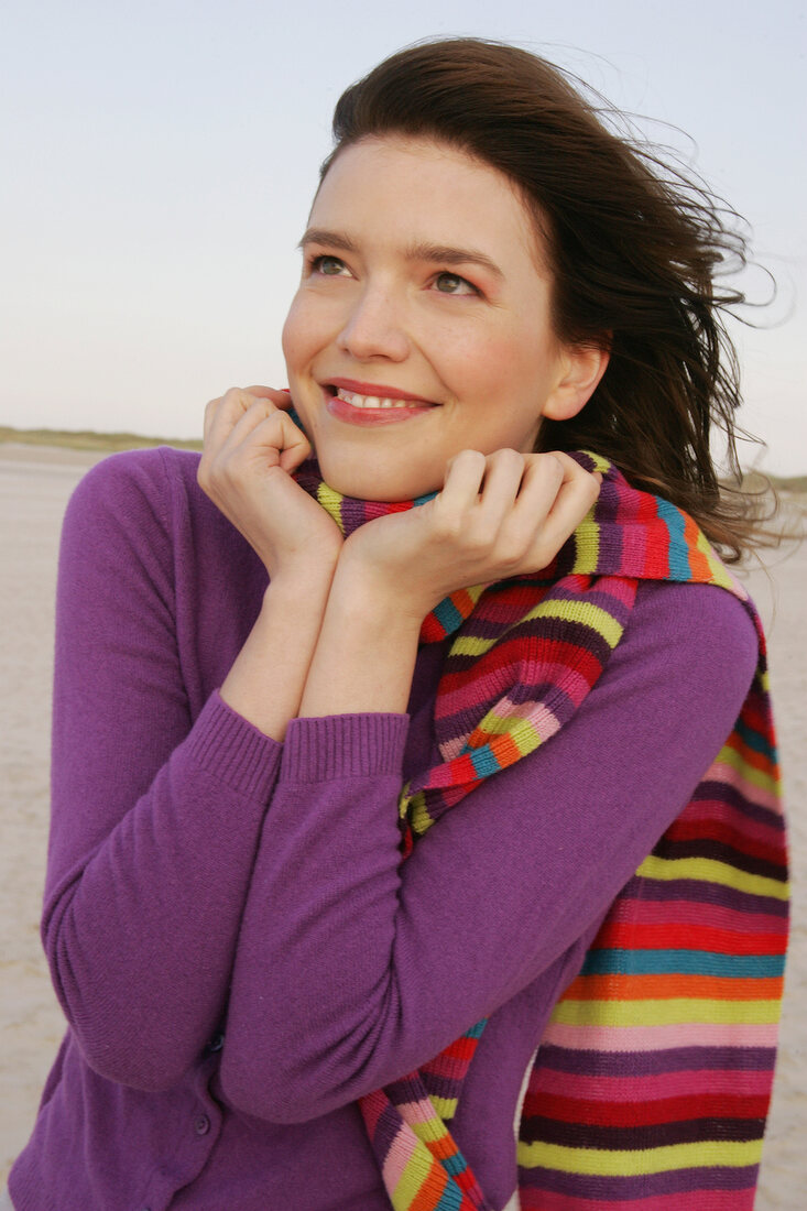 Pretty blonde woman wearing purple sweater and striped scarf standing on beach, smiling