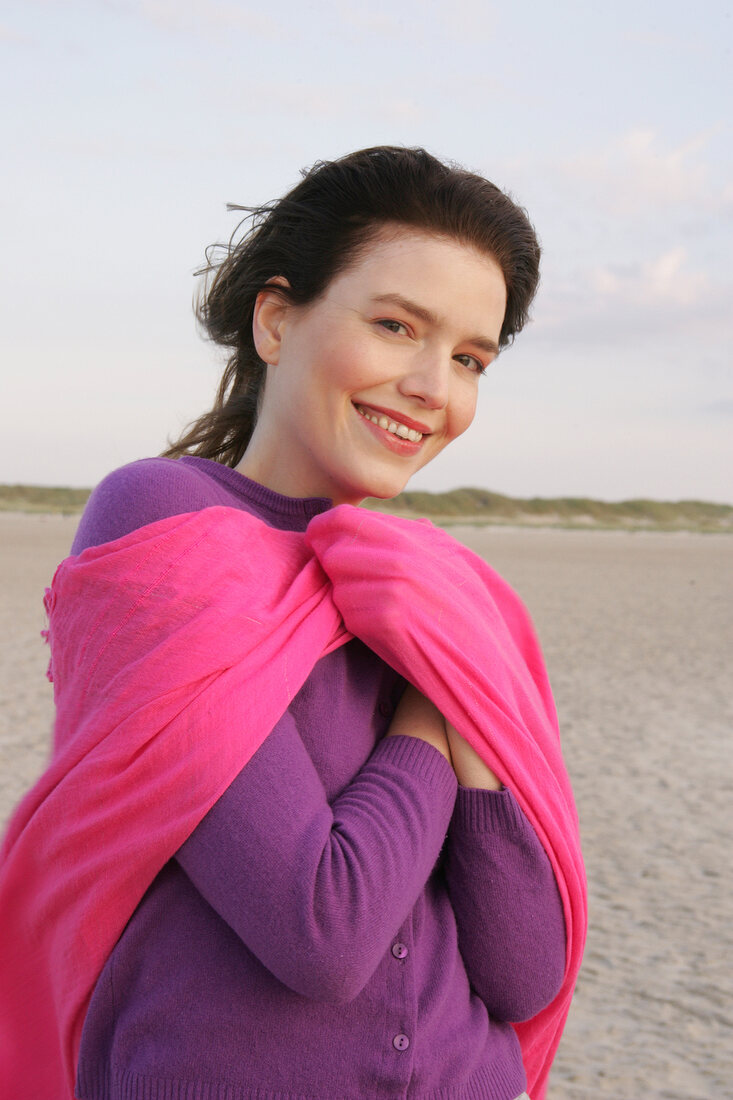 Portrait of pretty woman wearing purple sweater and pink shawl standing on beach, smiling