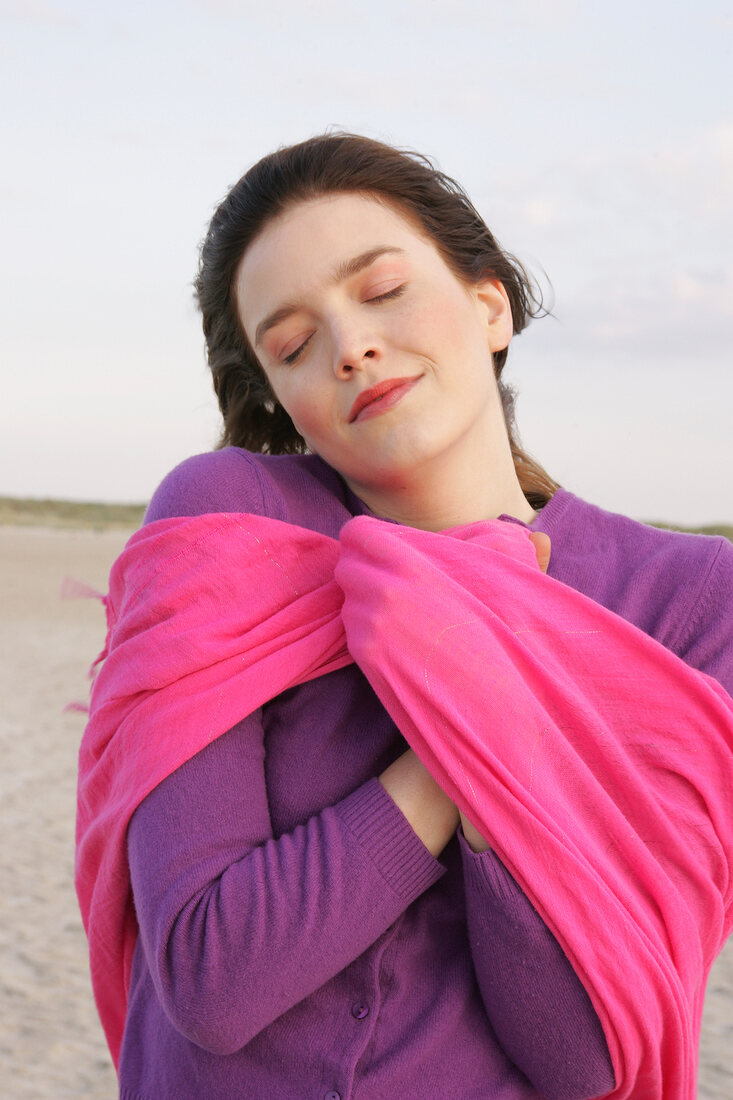 Portrait of pretty woman wearing purple sweater and pink shawl standing on beach, smiling