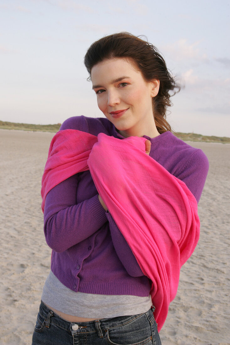 Portrait of pretty woman wearing purple sweater and pink shawl standing on beach, smiling