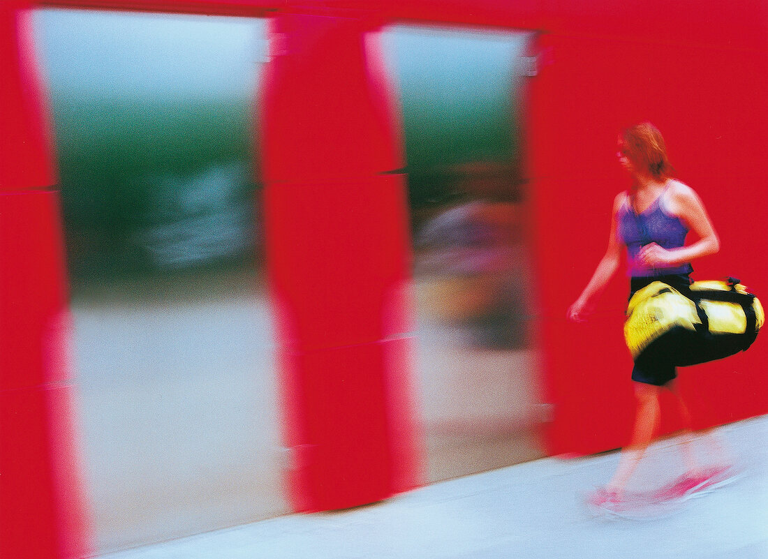 Frau im lila Top m. Sporttasche geht eilig vor roter Wand, verschwommen