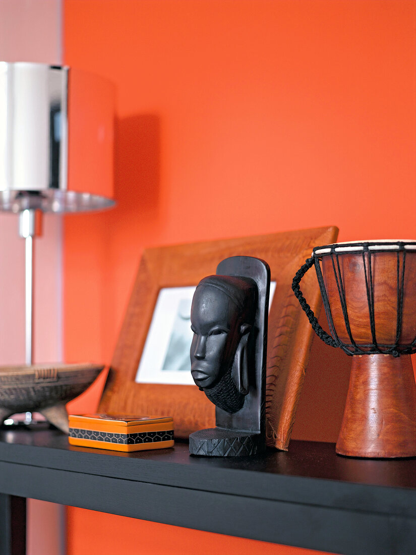 Mask statue with drum, picture frames and box on shelf against orange wall