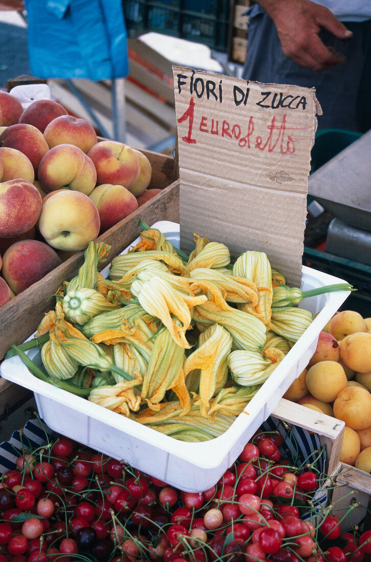 Zucchiniblüten, Äpfel, Kirschen und Orangen in Kisten auf einem Markt