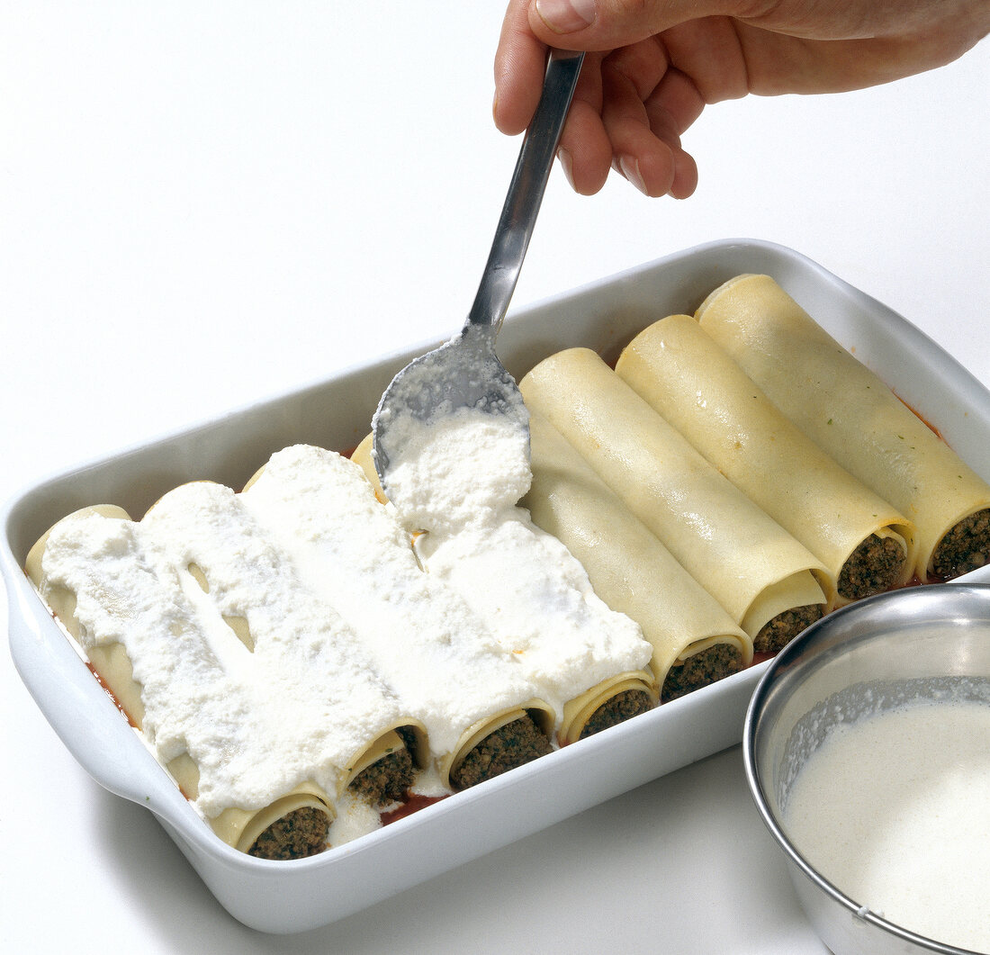 Cream being spread on stuffed pasta rolls while preparing cannelloni, step 3