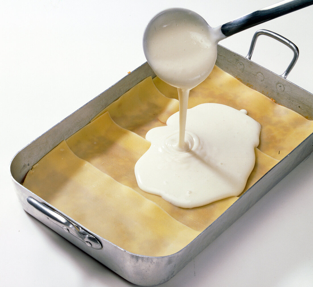 Bechamel sauce being poured on layers of dough sheet and meat sauce in baking tray