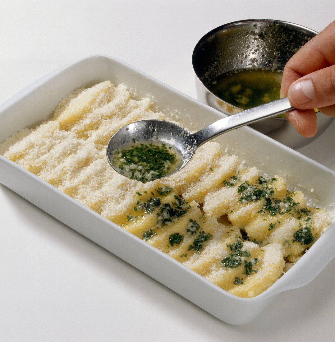 Herb butter being poured over gnocchi discs in baking dish