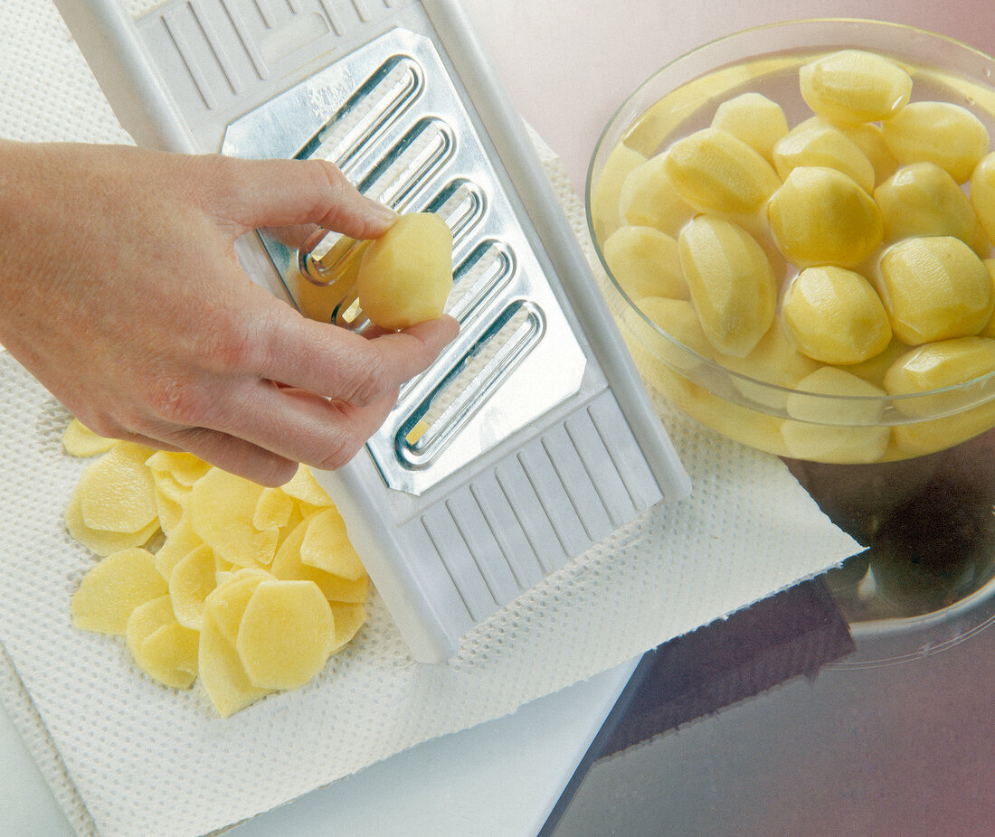 Peeled potatoes being grated in thin sliced on steel and plastic slicer with bowl