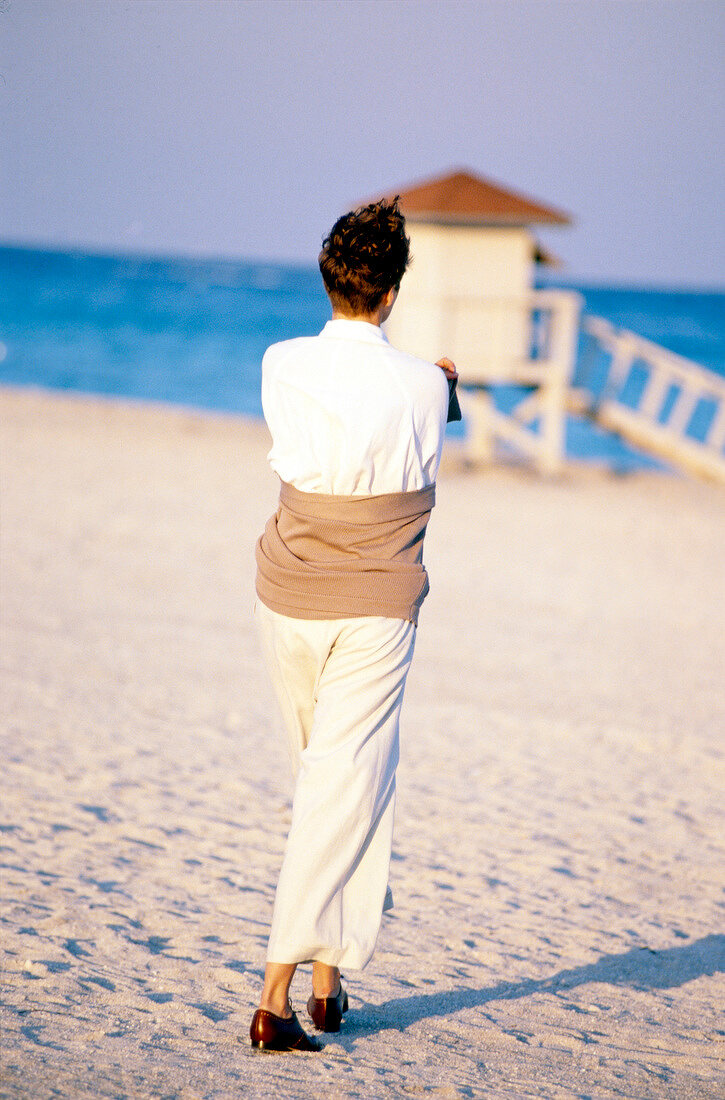 Frau mit Kurzhaarfrisur in Weiß geht am Strand spazieren, Rückansicht