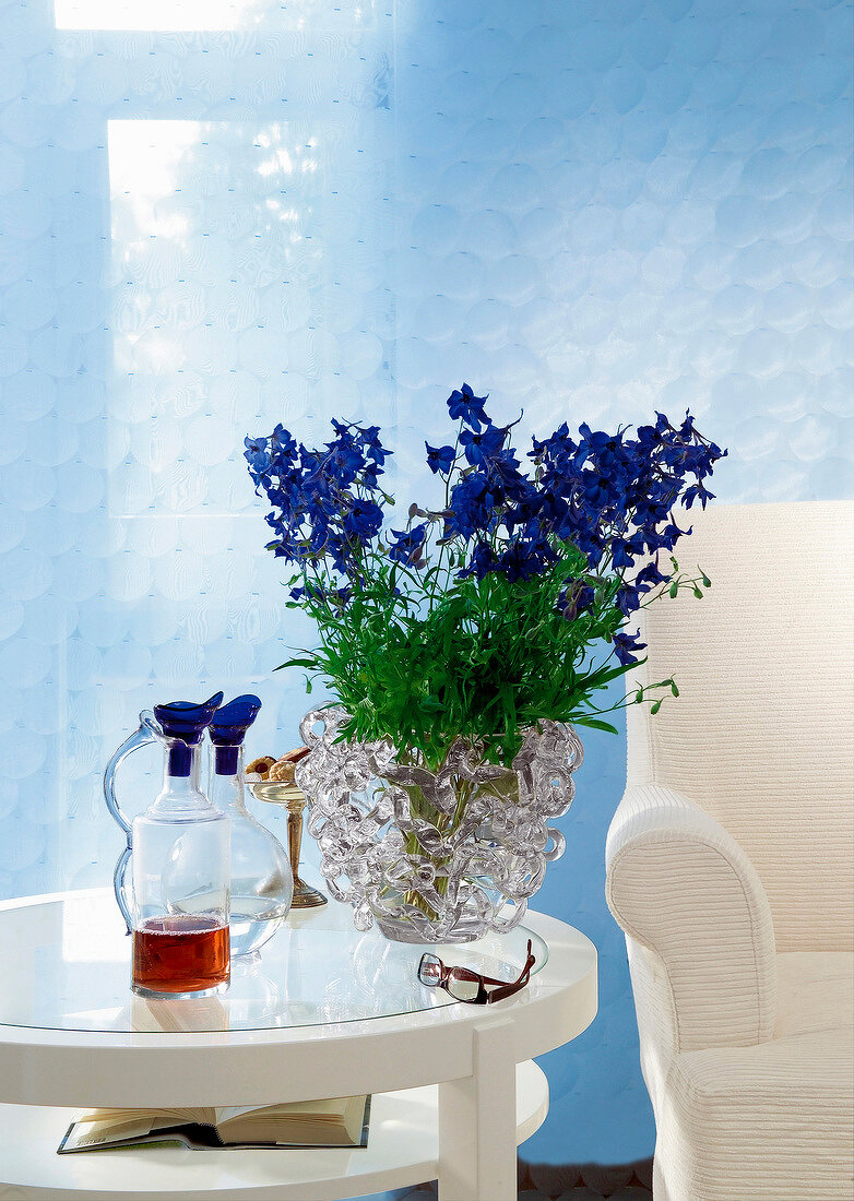 Glasvase mit lila Blumen auf Glastisch neben weißem Sessel