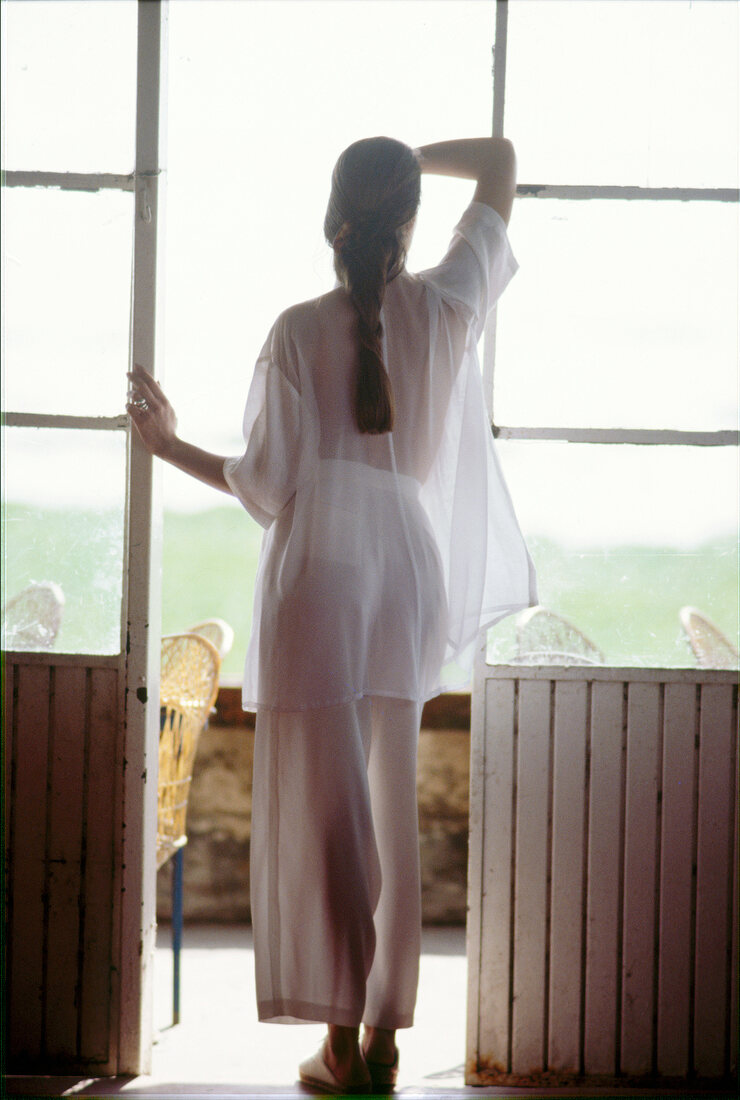 Brünette Frau mit Zopf in Weiß steht am Fenster, Rückansicht