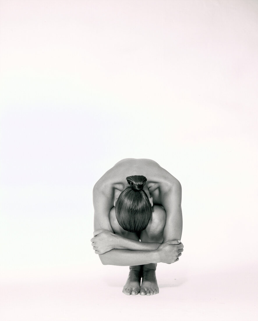Woman with hair bun crouching and hiding her face between knees against white background