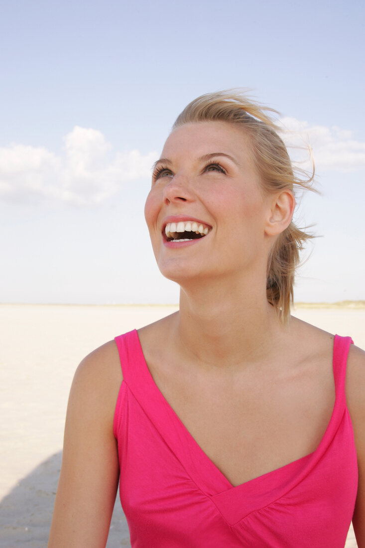 Happy blonde woman in pink dress looking up and smiling on beach