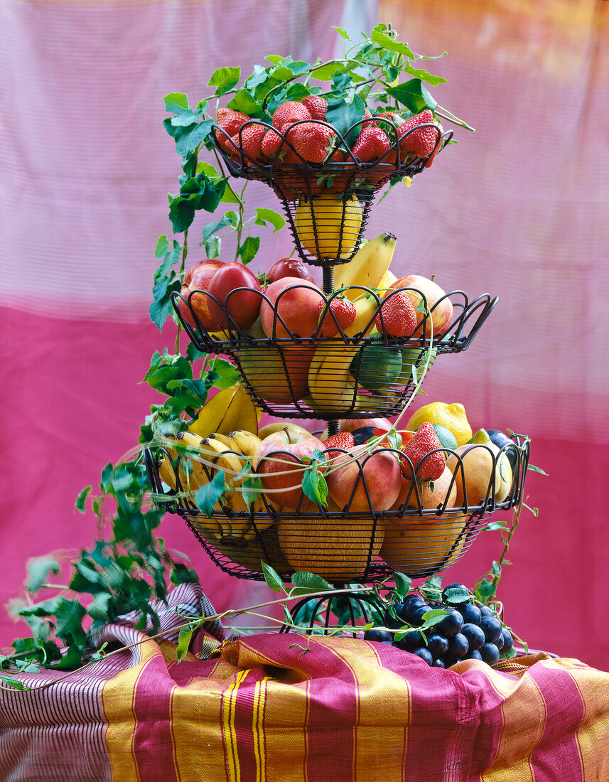 Different fresh fruits in tiered wire stand with tendrils on table