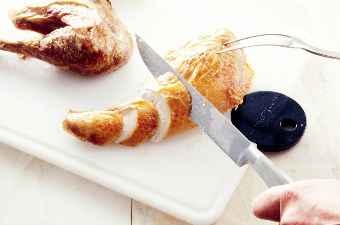 Baked chicken being cut into slices with knife and carving fork on chopping board, step 4