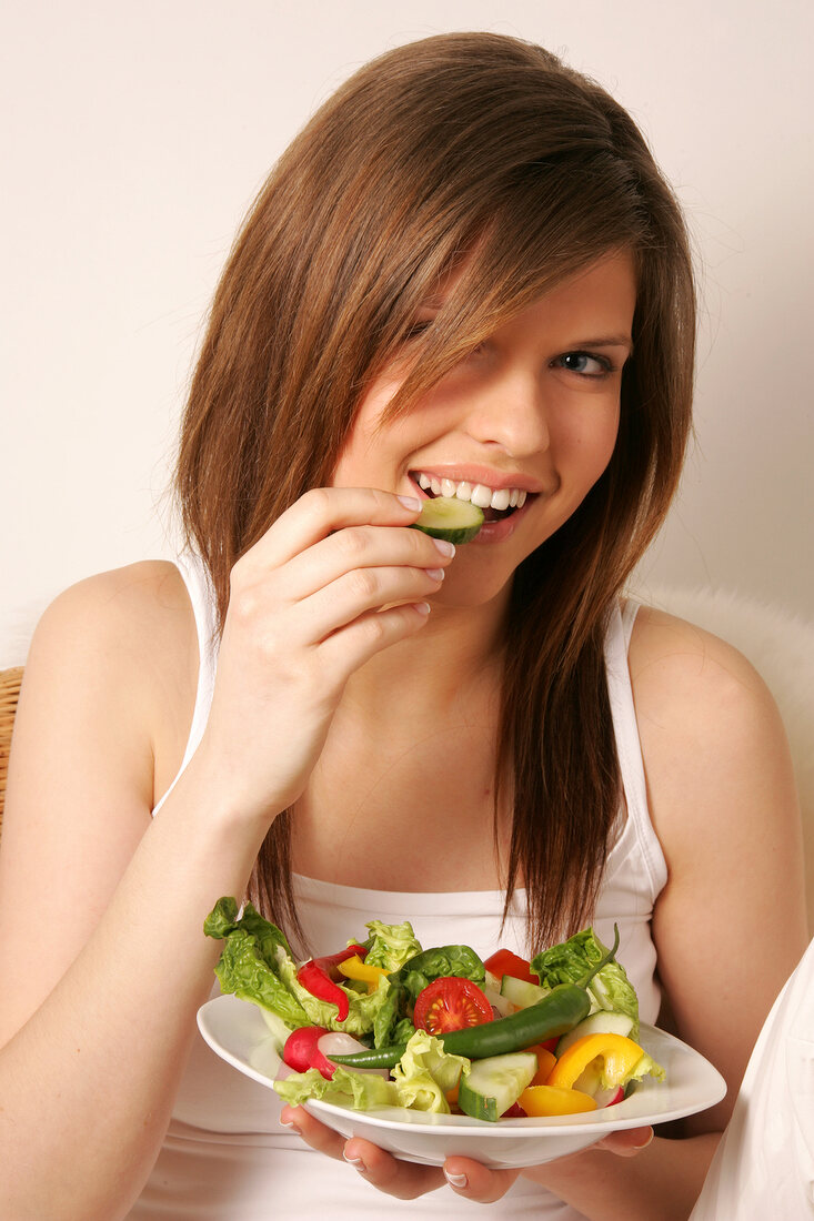 Portrait of woman with long hair holding bowl of salad and eating cucumber, smiling