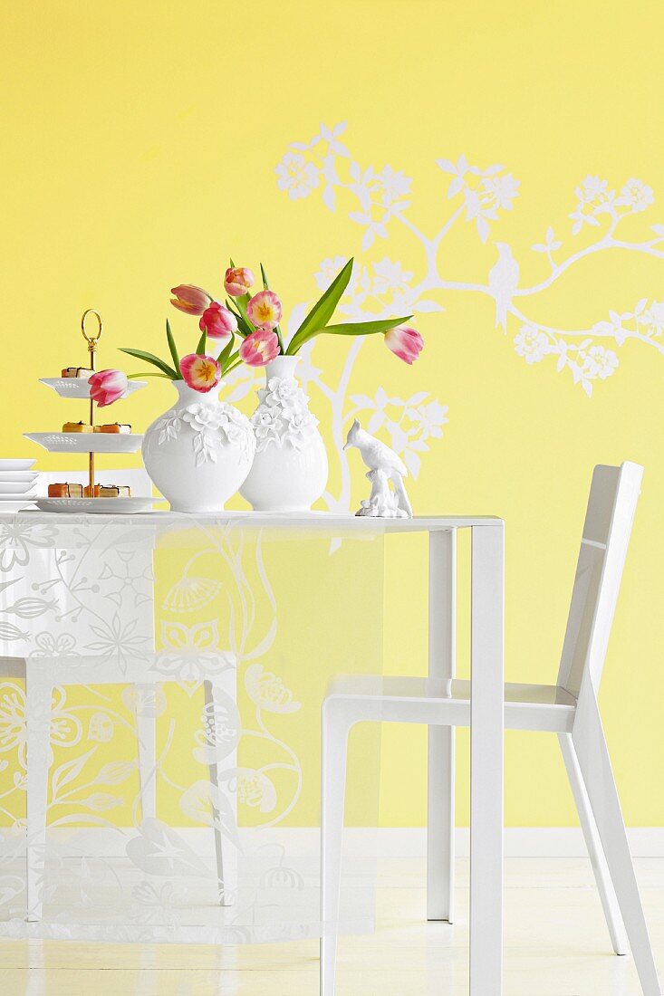 Two vases of tulips and cake stand of confectionary on white table in front of yellow wall