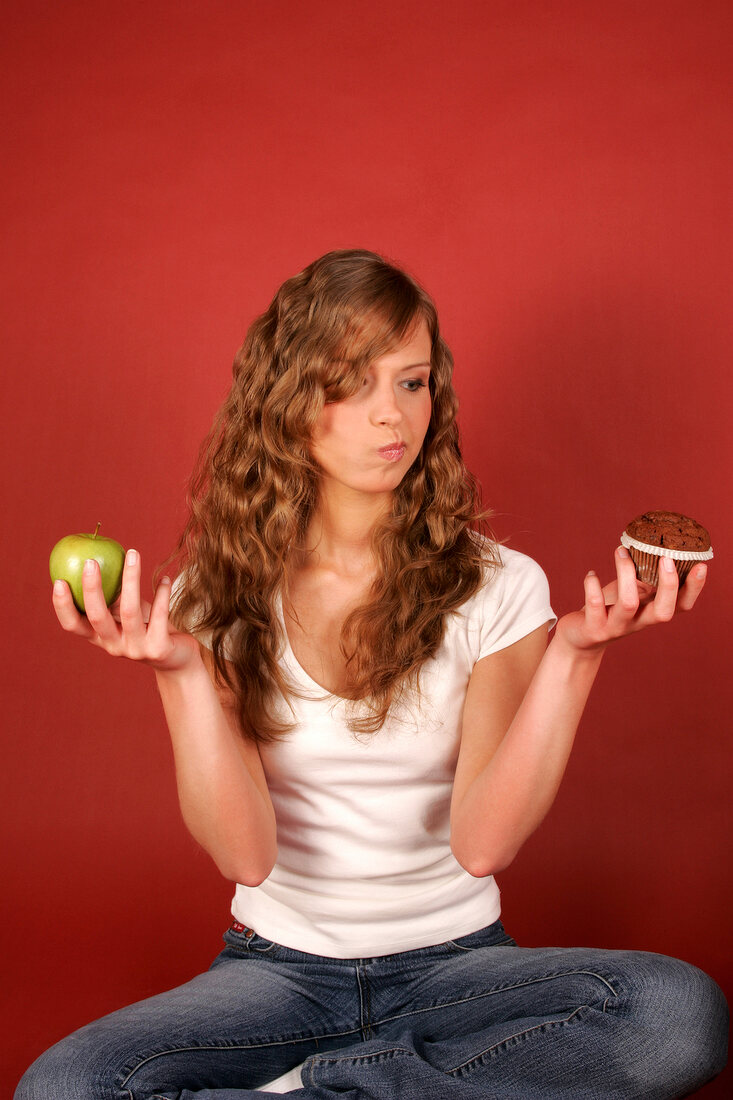 Pretty woman sitting cross legged holding green apple in one hand and muffin in other