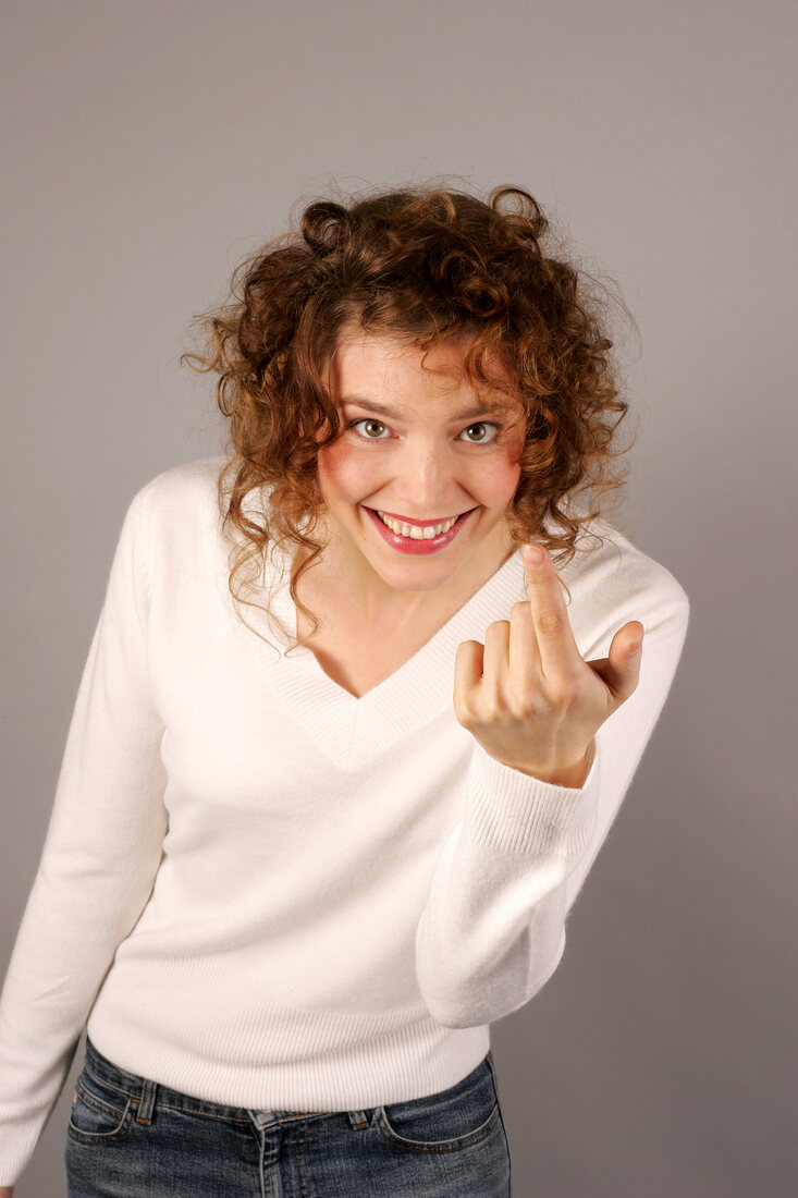 Cheerful young woman with curly hair wearing white sweater, gesturing with index finger