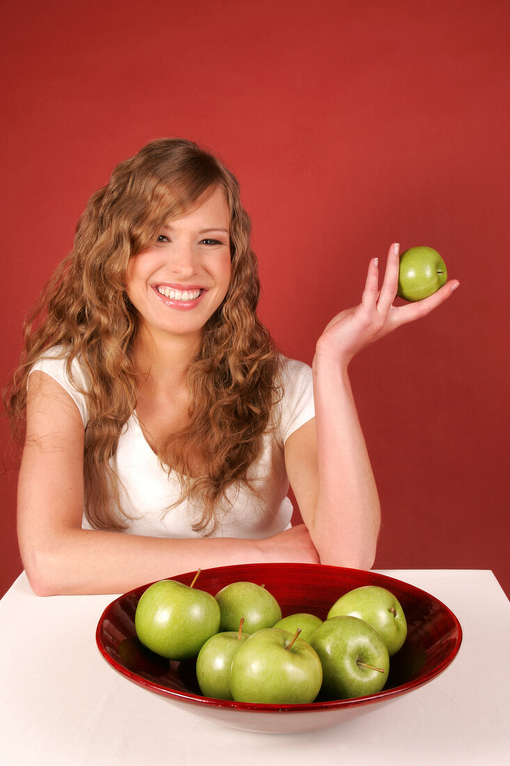 Happy woman sitting at table with bowl of green apples and holding apple in hand