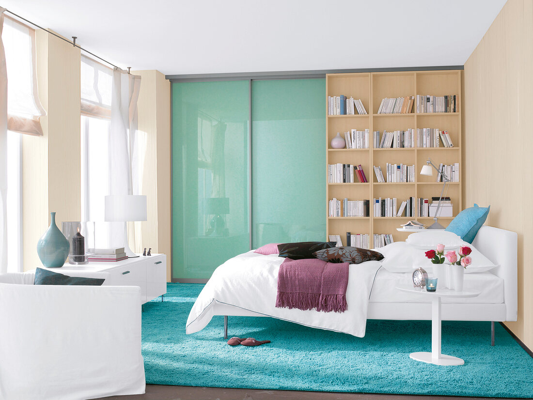 Bedroom in white and beige with blue double bed, bookcase and window