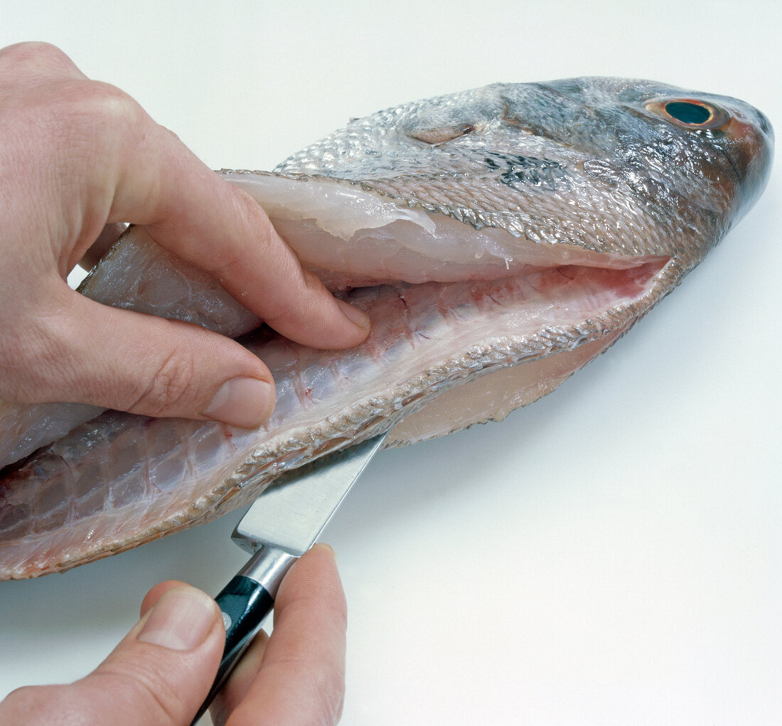 Close-up of hand cutting fish spine lengthwise on white background, step 2