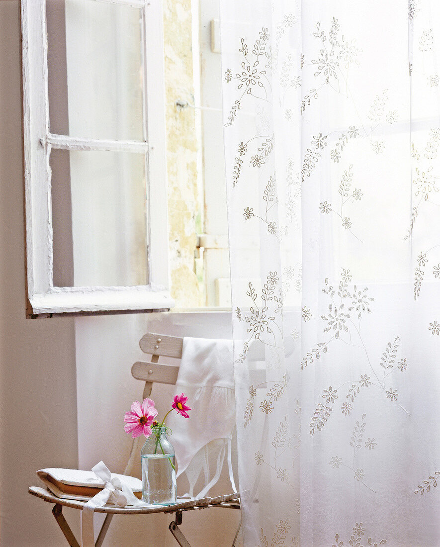 Transparent curtain, two flowers and handbag on chair beside open window