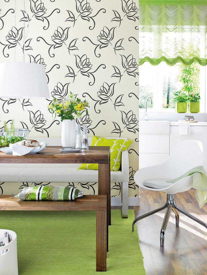 Laid wooden table and swivel chair in front of floral wallpaper with kitchen in background