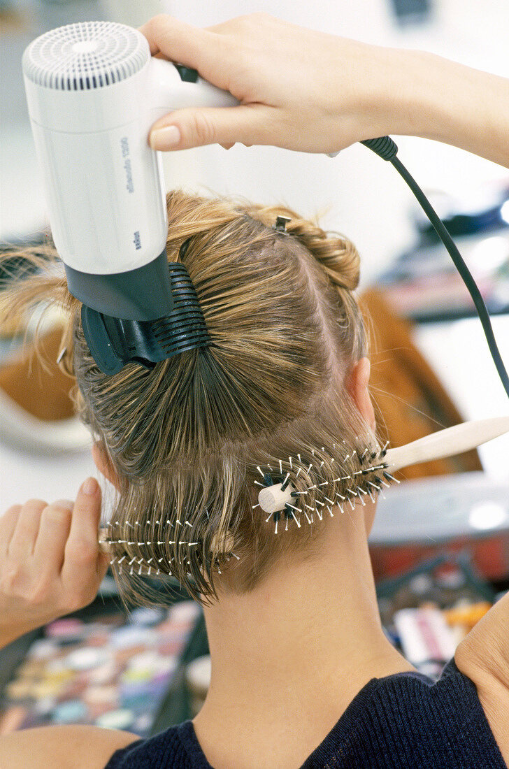 Rear view of blonde woman styling her hair with round brush and dryer