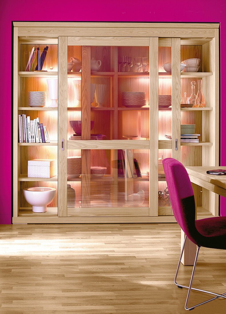 Illuminated oak wood cabinet with sliding glass doors in pink wall and chair on side