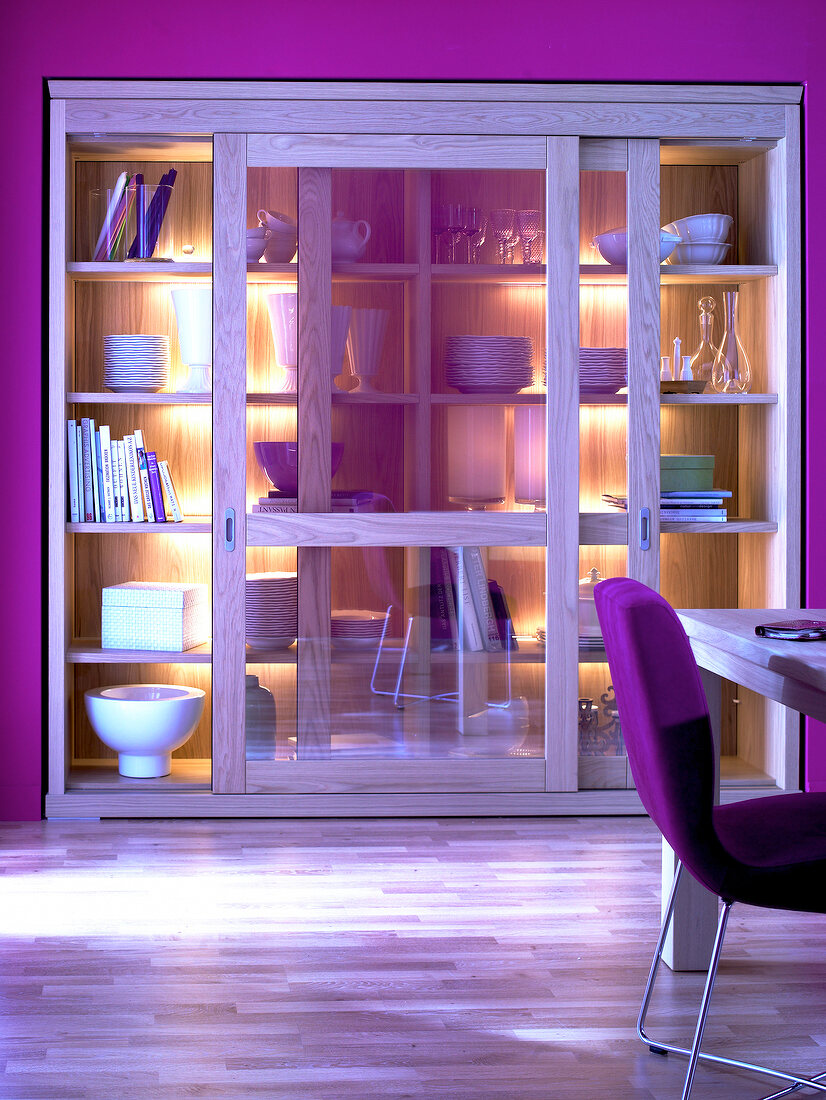 Illuminated oak wood cabinet with sliding glass doors in purple wall and chair on side