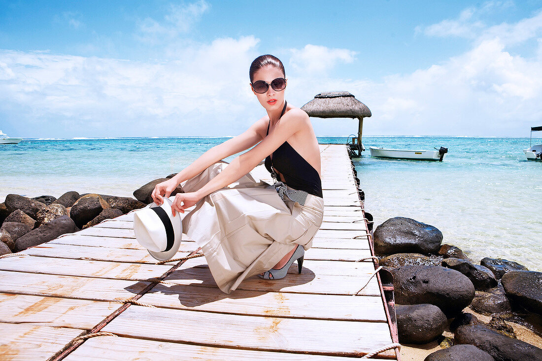 Fashionable woman wearing sunglasses holding hat and crouching on jetty