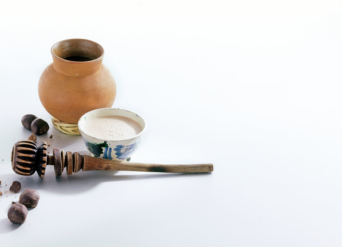 Cocoa powder in bowl with clay pot and wooden beater on white background