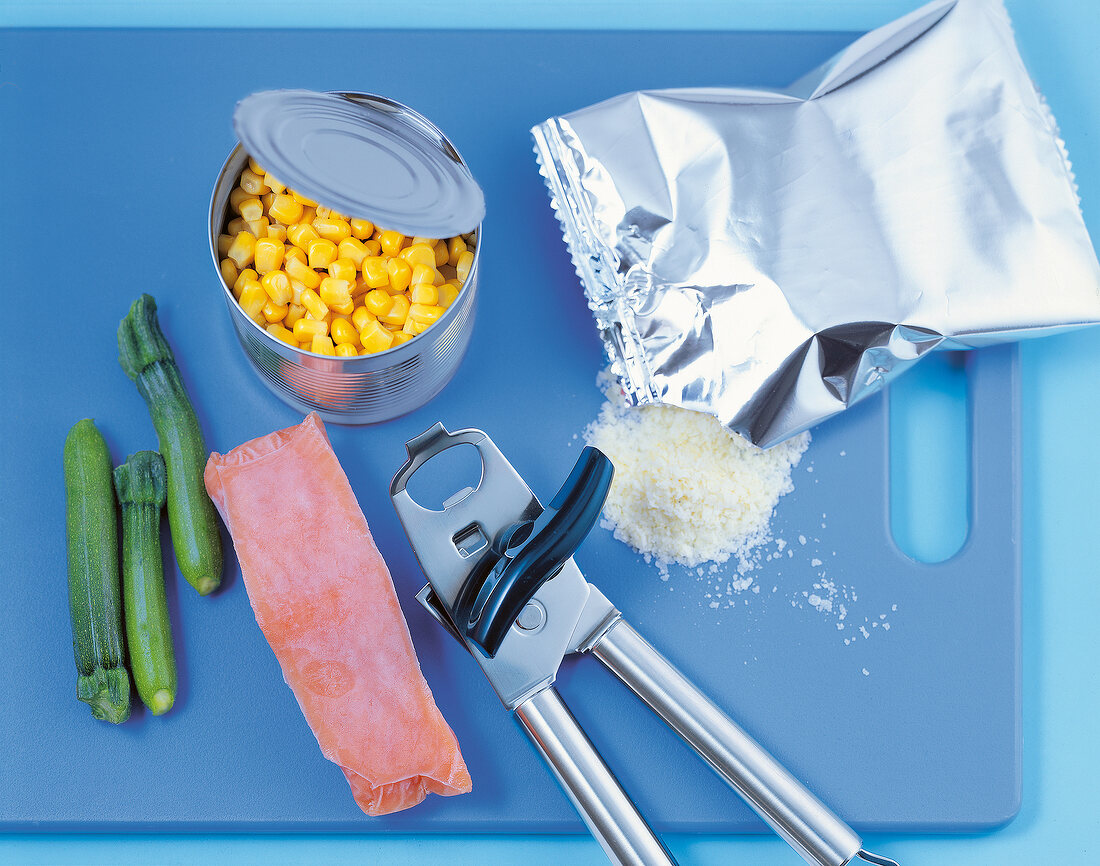 Canned corn, zucchini, frozen fish, can opener and powder on blue tray, overhead view
