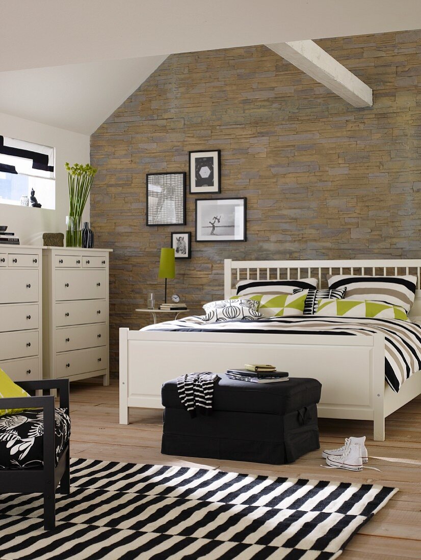 Weisses Doppelbett und Kommode im Schlafzimmer mit Bruchstein-Tapete; schwarz-weißer Teppich und Dachbalken